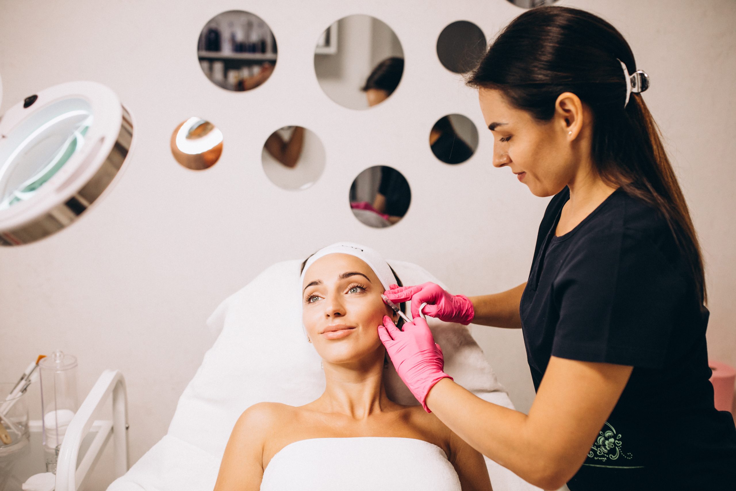 An image of a cosmetologist performing a beauty treatment on a woman at a modern beauty salon. The woman is lying comfortably on a white treatment bed with a relaxed expression, wearing a headband and a white towel. The cosmetologist, dressed in a black uniform with her hair neatly tied back, is seen wearing pink gloves and administering facial injections with precision. The salon's background features decorative circular mirrors arranged on the wall, creating a chic and contemporary ambiance, while professional skincare equipment is visible in the foreground.