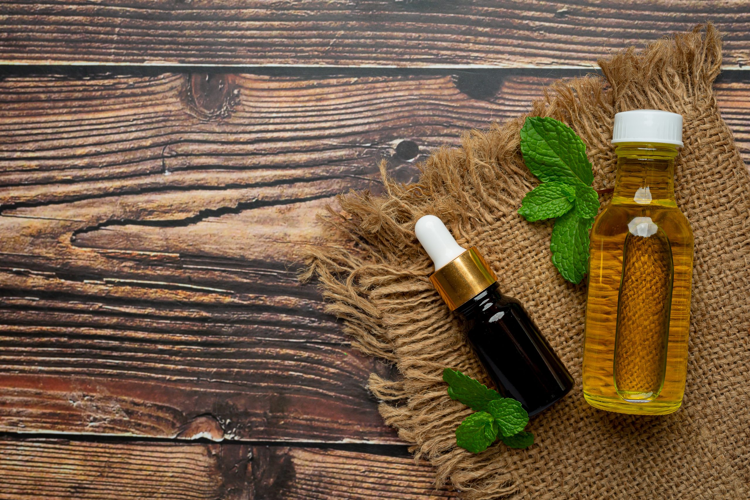 Two bottles of essential oils placed on a burlap cloth over a rustic wooden background, accompanied by fresh green peppermint leaves for a natural and organic aesthetic.