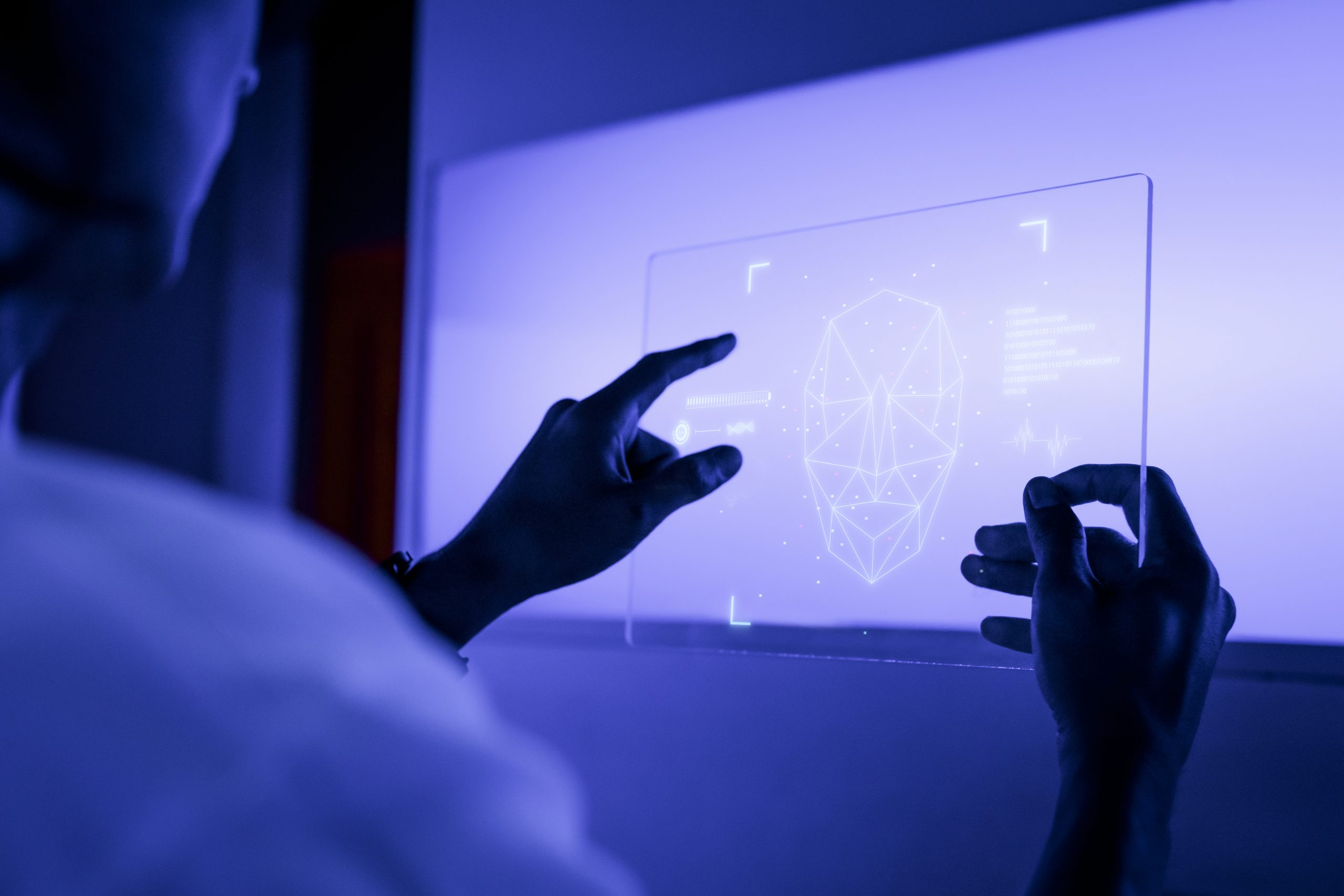 A futuristic image showcasing a person interacting with a transparent digital tablet screen. The screen displays a glowing facial recognition interface with a geometric outline of a face, data points, and other high-tech graphical elements. The scene is illuminated by a cool blue light, emphasizing the advanced technology and innovative design. The person's hands are seen manipulating the interface, suggesting a creative or analytical task in a modern, tech-driven environment.